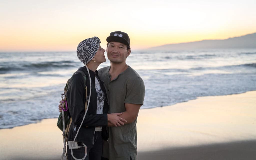 Smiling woman with long dark hair wearing a light pink blouse and black glasses standing outdoors in a modern city setting